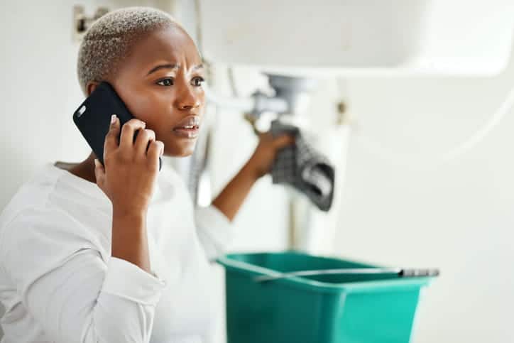 A woman on the phone with an emergency plumber as she holds up a rag to a leaking water heater. Underneath is a bucket to collect excess water.