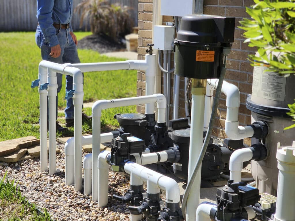 Swimming pool plumbing pipes and system at residence with plumber working in background in the yard.